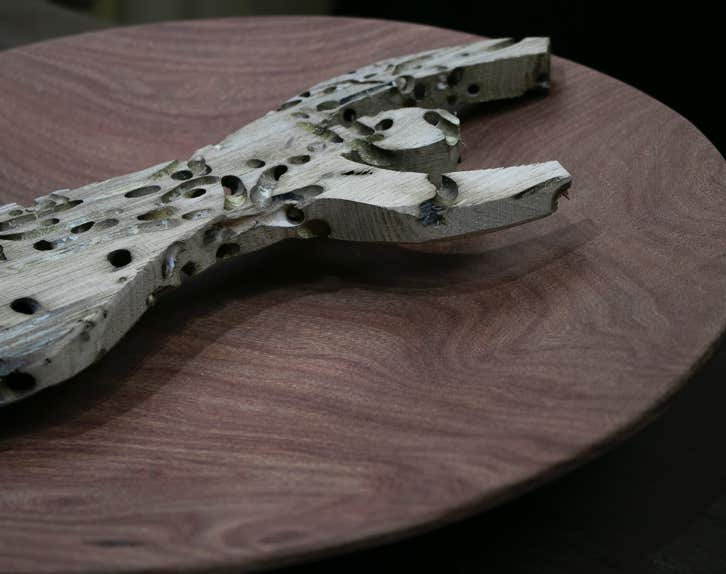 Scultura in legno riciclato raffigurante una donna che alza le mani distesa su di un piatto di legno.